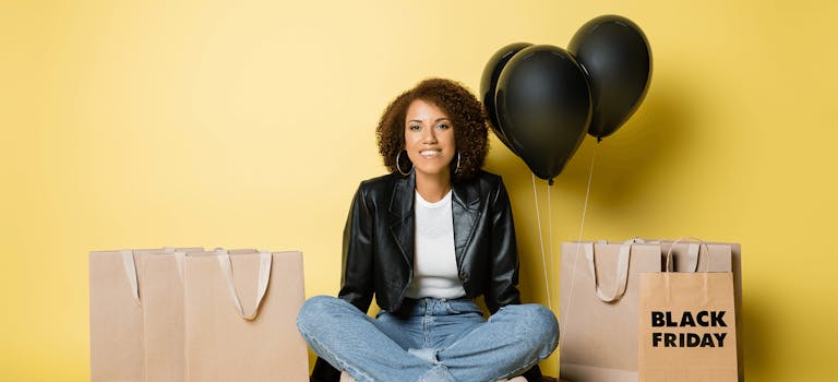 Woman sitting cross-legged against yellow wall with paper bags and black balloons around her.