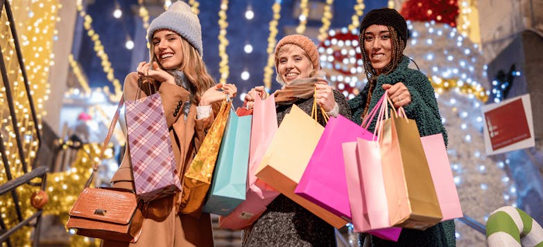 Three women in woolly hats and coats holding colourful shopping bags against Christmas lights background