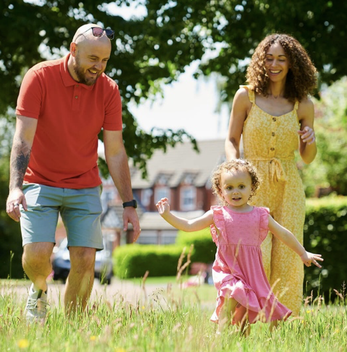 A family walking through long grass in a green open space, with houses and hedgerows beyond