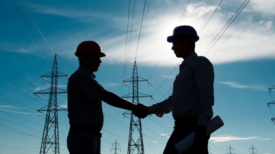 Inżynierowie elektrotechnicy ściskają sobie ręce na tle kabli i linii wysokiego napięcia