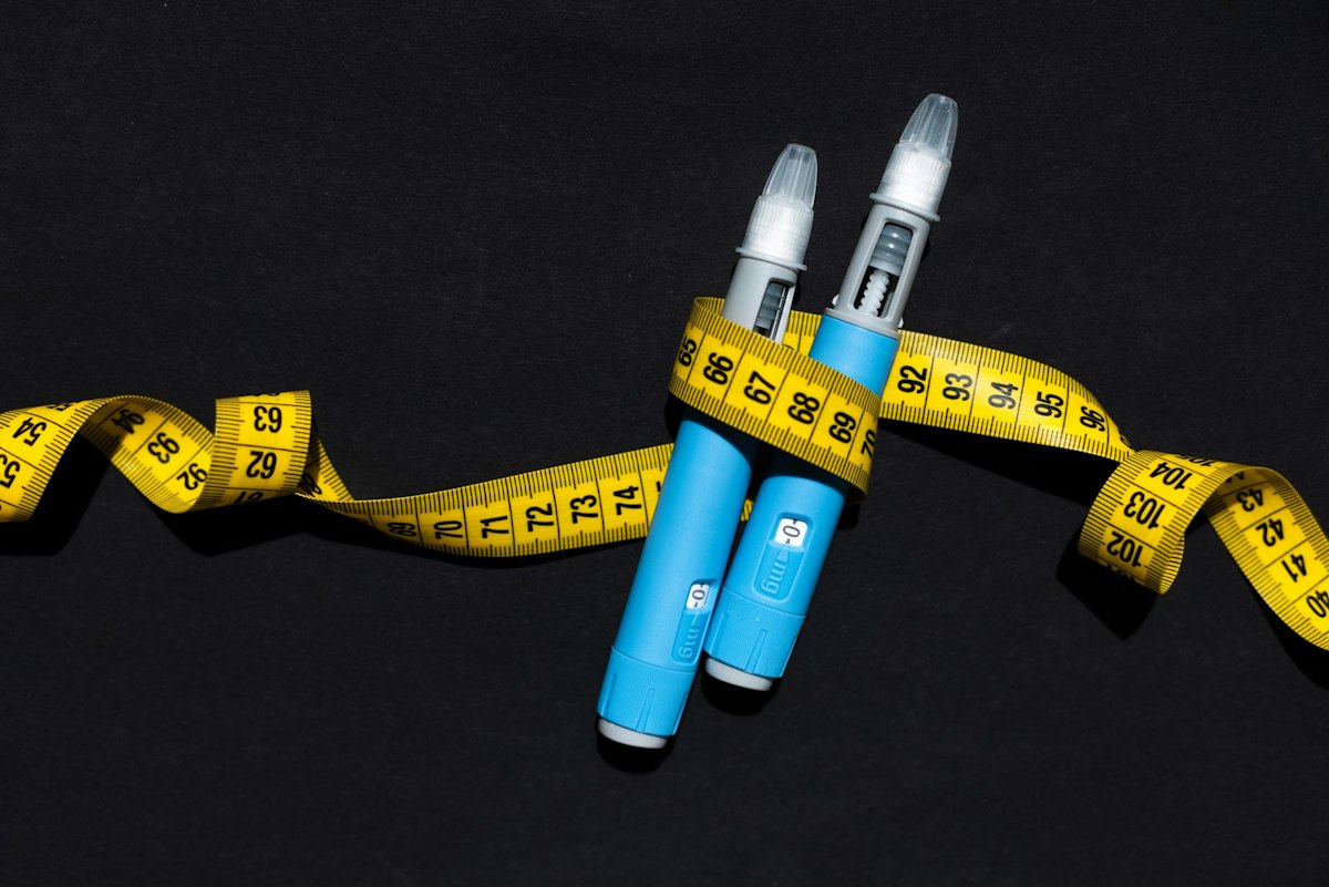 Two blue GLP 1 weight loss injection pens wrapped in a yellow measuring tape on a black background, representing medical weight management.