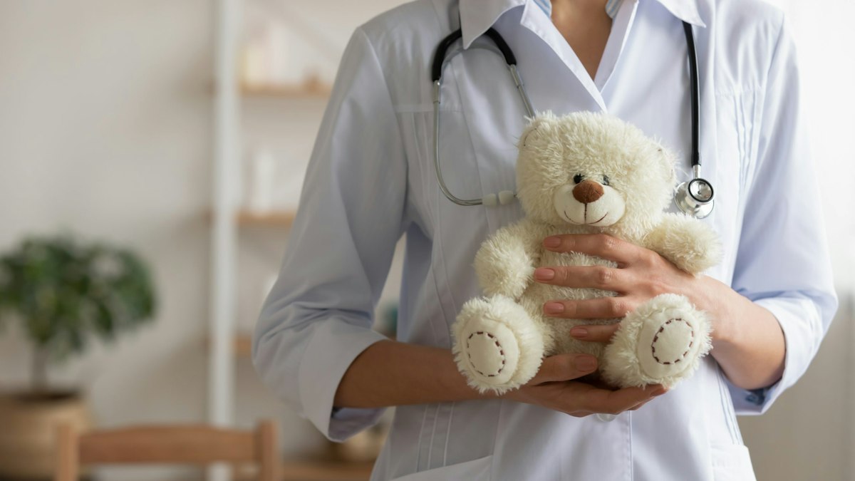 Female pediatrician holding a teddy bear while speaking with a young patient in a warm clinical setting at Geneva.