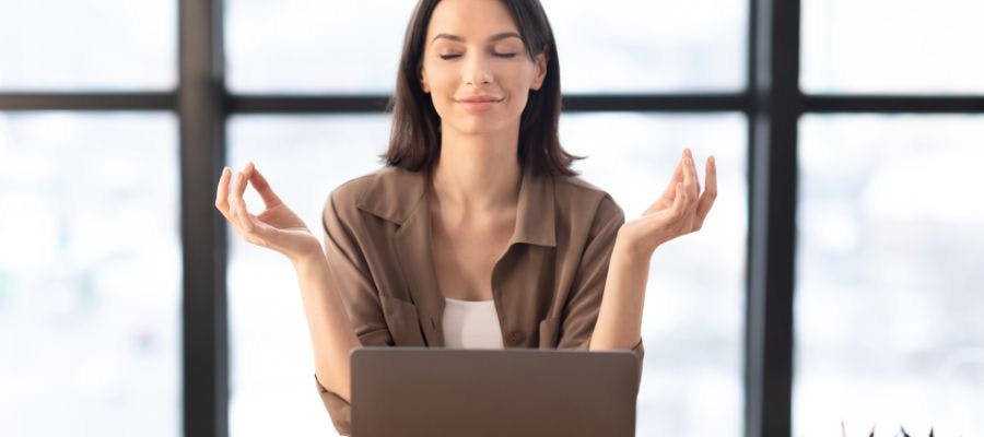 A woman is sitting at a desk, holding her hands in front of her, and smiling. She is wearing a brown shirt and has her hands in front of her. She is looking at a laptop computer that is open in front of her. The background shows a window with a view of a cityscape. A woman is sitting at a desk, holding her hands in front of her, and smiling. She is wearing a brown shirt and has her hands in front of her. She is looking at a laptop computer that is open in front of her. The background shows a window with a view of a cityscape.