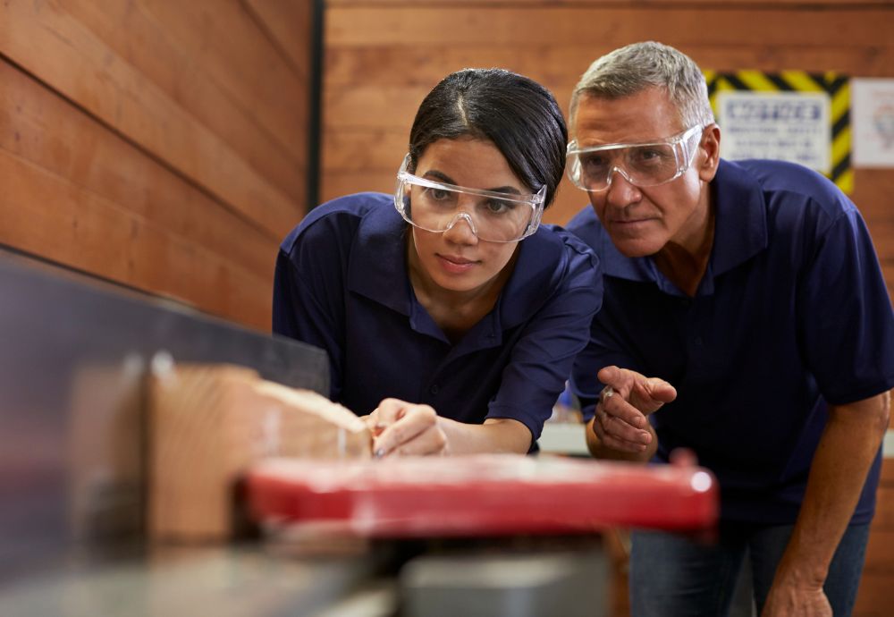 A person showing an apprentice how to sand wood