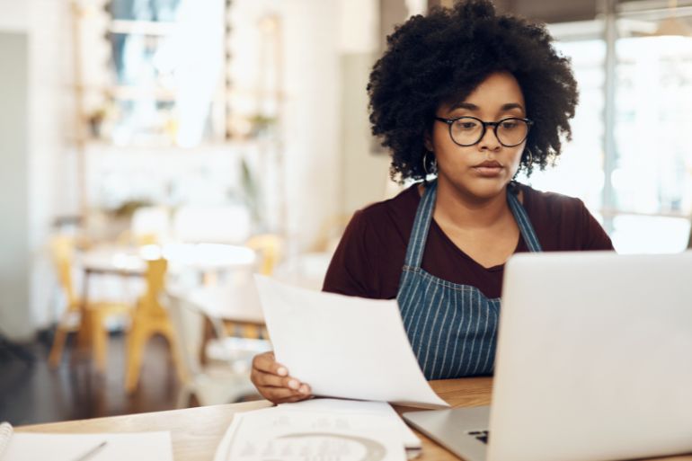 A woman in glasses and an apron sits at a table with papers and a laptop, working on payroll processing