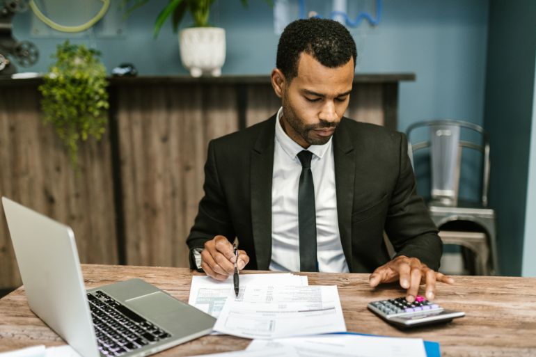 A man in a suit focused on his laptop and paperwork, calculating payroll