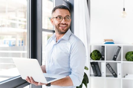What is HR software man happily holds a laptop in an office. Using cloud-based HR software on his .