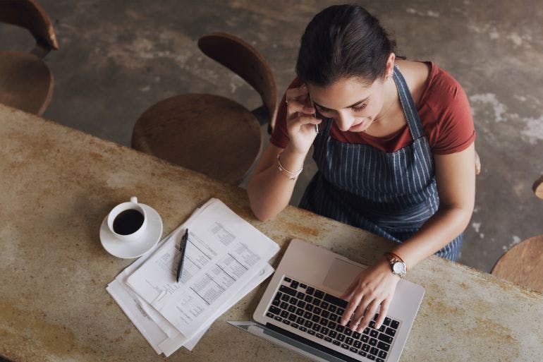 Female business owner itting at a table with papers and coffee, working on her laptop utilizing essential payroll features