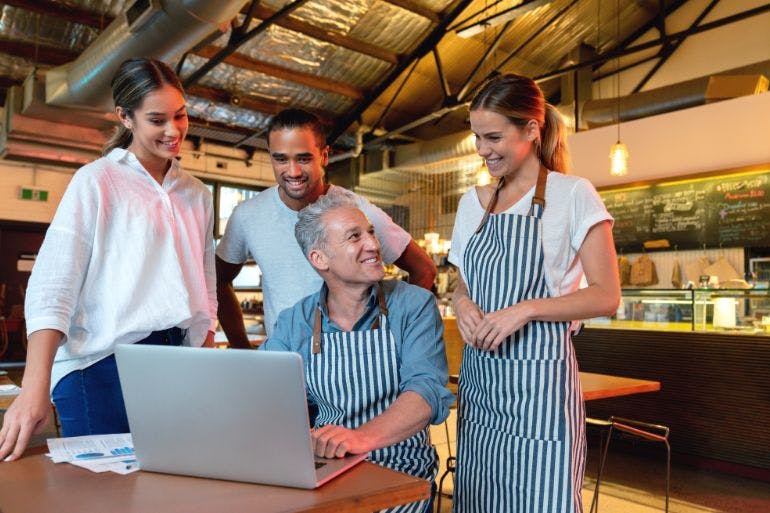 Rota planning Business owner and employees in aprons standing around a laptop, discussing rota planning.