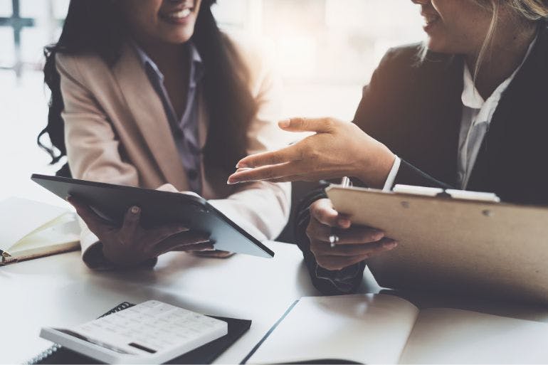 Two businesswomen chatting about different types of payroll software at a table with a tablet.