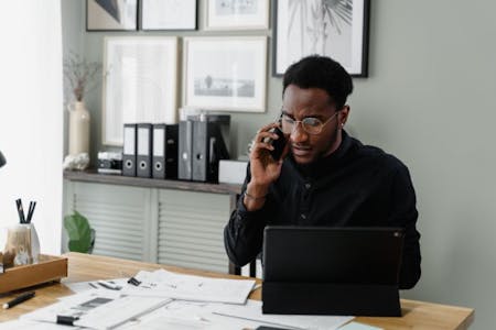 A man in glasses sits at a desk with a laptop and a phone, researching types of payroll software.
