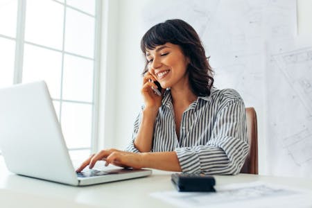 Female business owner on phone at desk with laptop, reviewing payroll records.