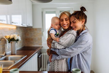 Shared parental leave Same sex parents holding their baby in a kitchen.