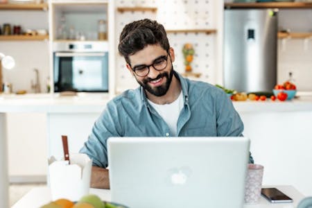 Remote onboarding A man with a beard and glasses sitting at a table with a laptop, engaging in remote onboarding.