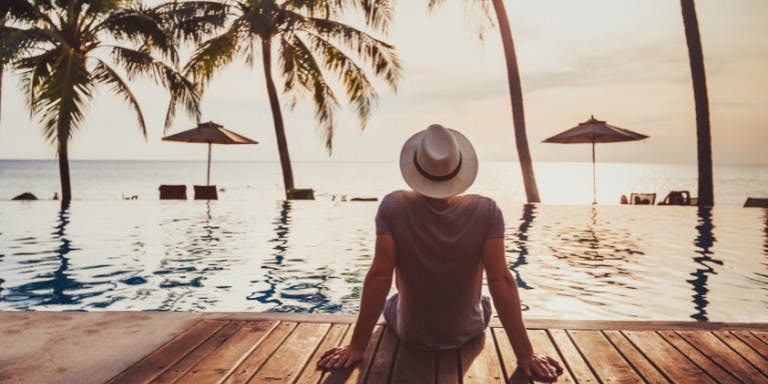Man wearing a hat sits by the pool, with palm trees in the background, symbolizing a well-deserved vacation from work.