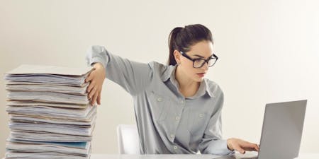 Woman in glasses pushing a stack of documents off a desk and looking hapy towards a laptop with online document storage.