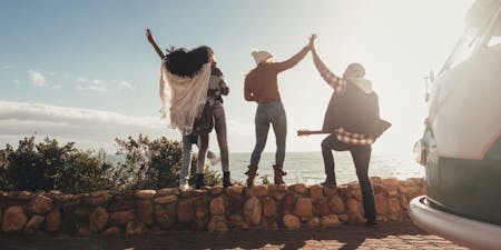 Annual Leave Four people pose with hands raised beside a camper van, embodying the spirit of adventure during their annual leave.