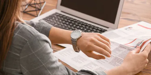 Business owner looking at watch at a desk with a newspaper and laptop.