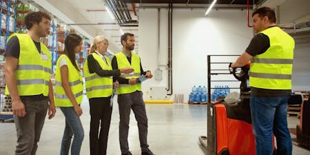 Health and safety management system Group of employees in a warehouse observing health and safety procedures for operating a forklift.