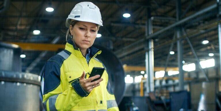 Risk Assesments Woman in a hard hat and safety vest uses her smartphone to perform risk assessments in a warehouse.