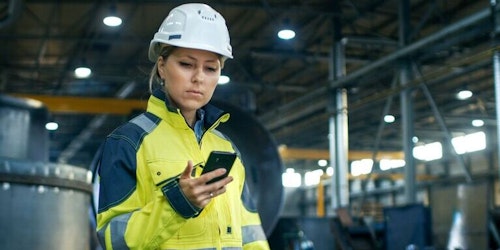 Woman in a hard hat and safety vest uses her smartphone to perform risk assessments in a warehouse.
