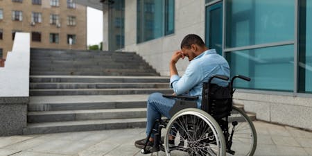 Types of discrimination Man in a wheelchair in front of steps into work building, highlighting the challenges of workplace discrimination faced by individuals with disabilities.