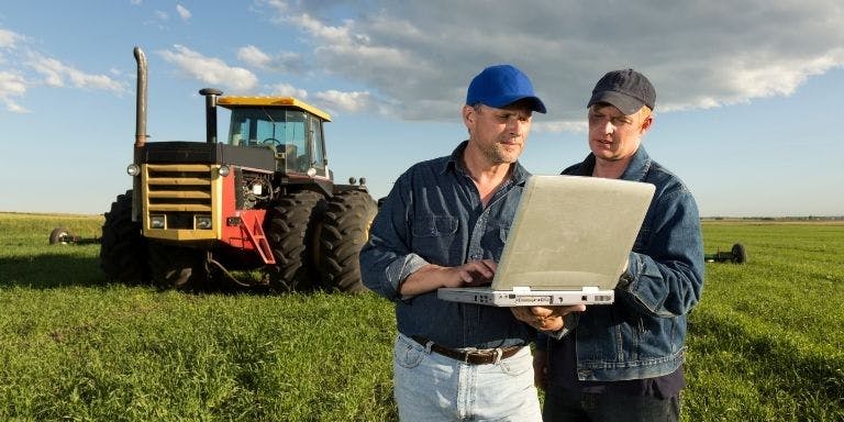 Two men stood reading from a laptop in a field, they are stood in front of a tractor