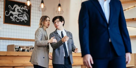 Two business professionals in suits pointing at a co-worker and laughing, showcasing harassment at work.