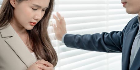 Man and woman stand in front of blinds, their body language suggesting an uncomfortable situation involving harassment.