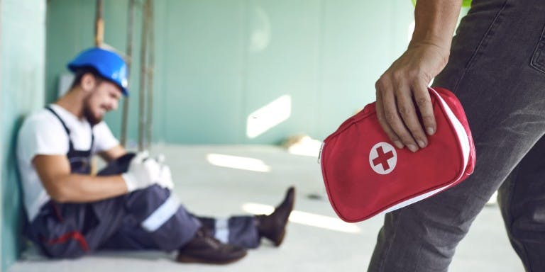 First aid at work An injured person on the floor and another bringing them a first aid kit, highlighting the importance of first aid readiness at work.