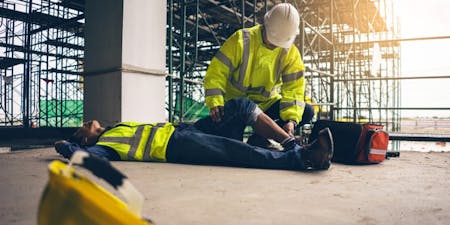 First aid at work man in a hard hat and safety vest is positioned on the ground, highlighting a first aid scenario at a construction site.