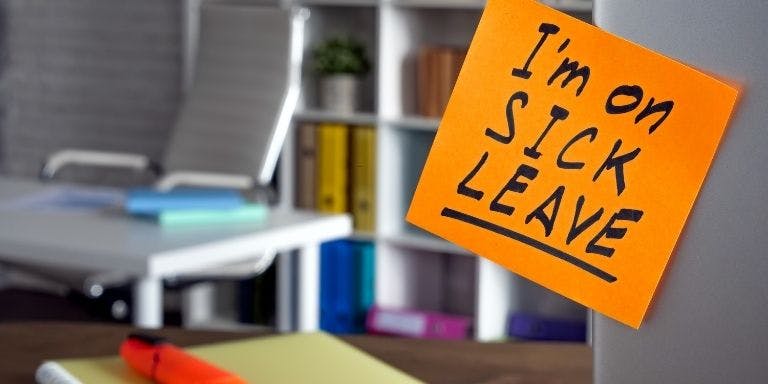 Notepad stuck on back of laptop saying "I'm on sick leave" in office