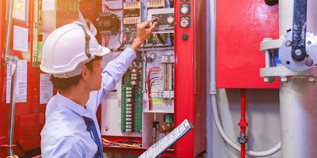 Fire safety at work Worker wearing a hard hat and safety vest is engaged in maintenance of a fire alarm panel for workplace safety.