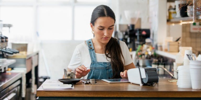 Allocation of tips in the workplace A woman in a blue overalls is seated at a coffee shop counter, holding a glass of coffee and examining it. The counter is cluttered with various objects including a coffee machine, a coffee cup, and a coffee maker. The woman appears to be in the process of making a purchase.