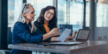 Two women seated at a table, focused on a laptop and papers while discussing a working hours agreement.