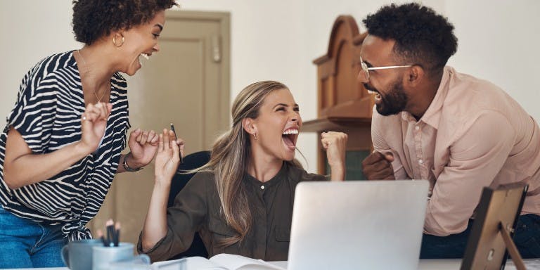 Three employees in front of a desk at work happy and celebrating great pay and benefits.