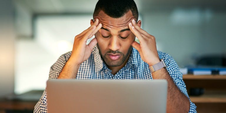 A man sat at a desk infront of a laptop appearing stressed
