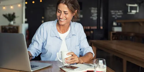 Female business owner at a table, working on a laptop with a coffee cup beside her, focused on managing her timesheets.