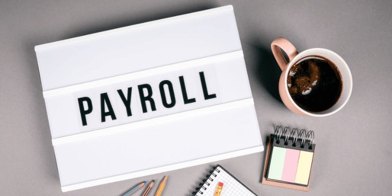 A folder reading "Payroll" from a birds eye view, next to a mug of coffee and some stationary.