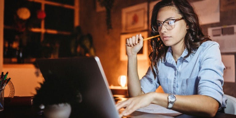 A woman wearing glasses diligently works on her laptop during a night shift, focused on her tasks.