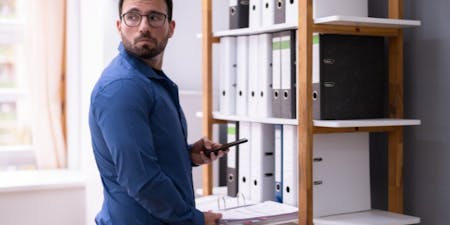 Gross misconduct at work Male employee taking pictures of confidential files on a phone in an office.