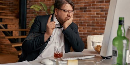 Gross misconduct at work Male employee drunk at his desk drinking a glass of whiskey with an empty bottle.