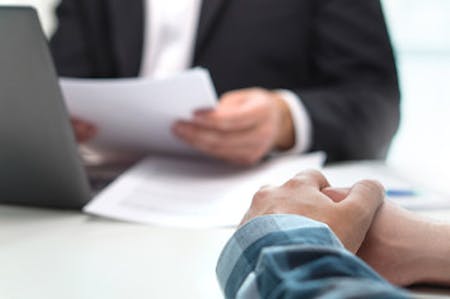 A shot of an employees hands, opposite over a desk to an employer holding some documents next to their laptop.