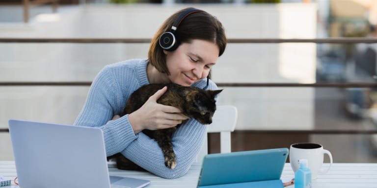 Female employee wearing headphones and using a laptop while holding a cat, illustrating flexible working arrangements at home.