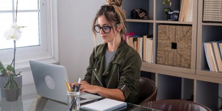Woman wearing glasses sits at a desk, focused on her laptop while writing performance objectives for work.