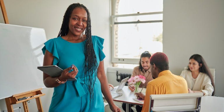 Woman in a blue dress stands confidently before a group of engaged individuals, symbolising development and collaboration.