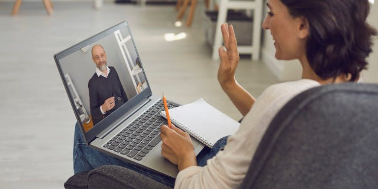 Woman seated in a chair, focused on her laptop, with a man visible on the screen, engaged in a work learning session.