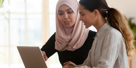 Developing employees through mentoring Two women collaborate over a laptop, engaged in a mentoring session at work.