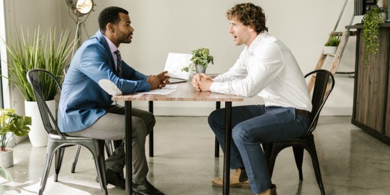 Male manager and male employee in a performance review meeting seated at a table in an office.