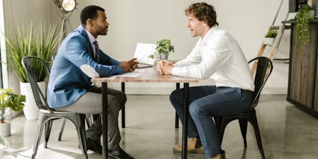 Performance review stage Male manager and male employee in a performance review meeting seated at a table in an office.
